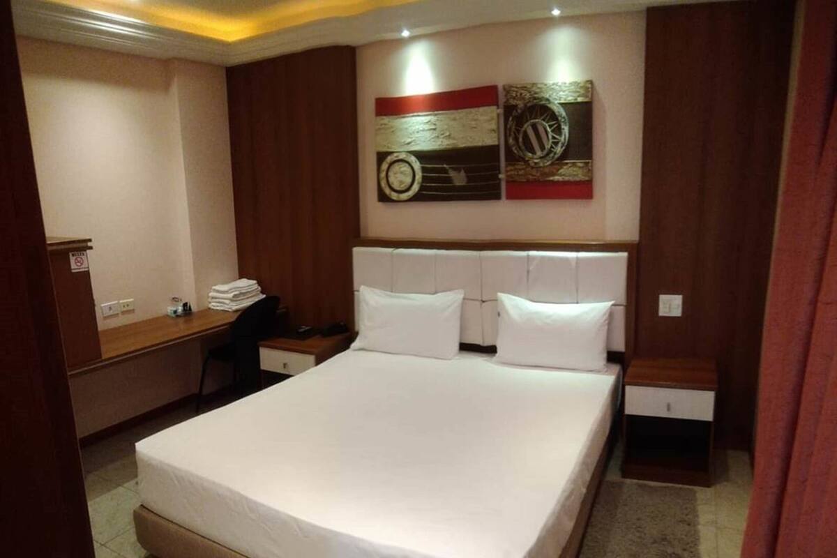 Ein Hotelzimmer mit einem großen Bett, einem Schreibtisch und einem Sessel. An der Wand hängt eine Uhr und ein gerahmtes Bild über dem Bett.