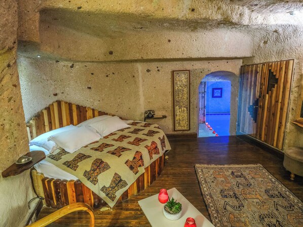 A rustic room with a wooden bed, a small table with a plant, a patterned rug, and a view through an archway. - Cappadocia Minia Cave Hotel (Ürgüp)