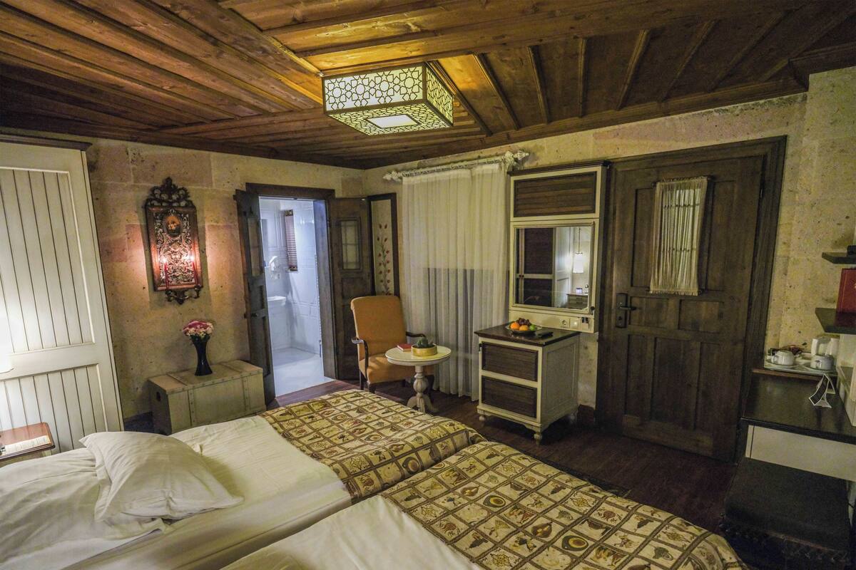 Habitación de hotel con dos camas, una mesita con fruta, una silla, un espejo y techo de madera.
