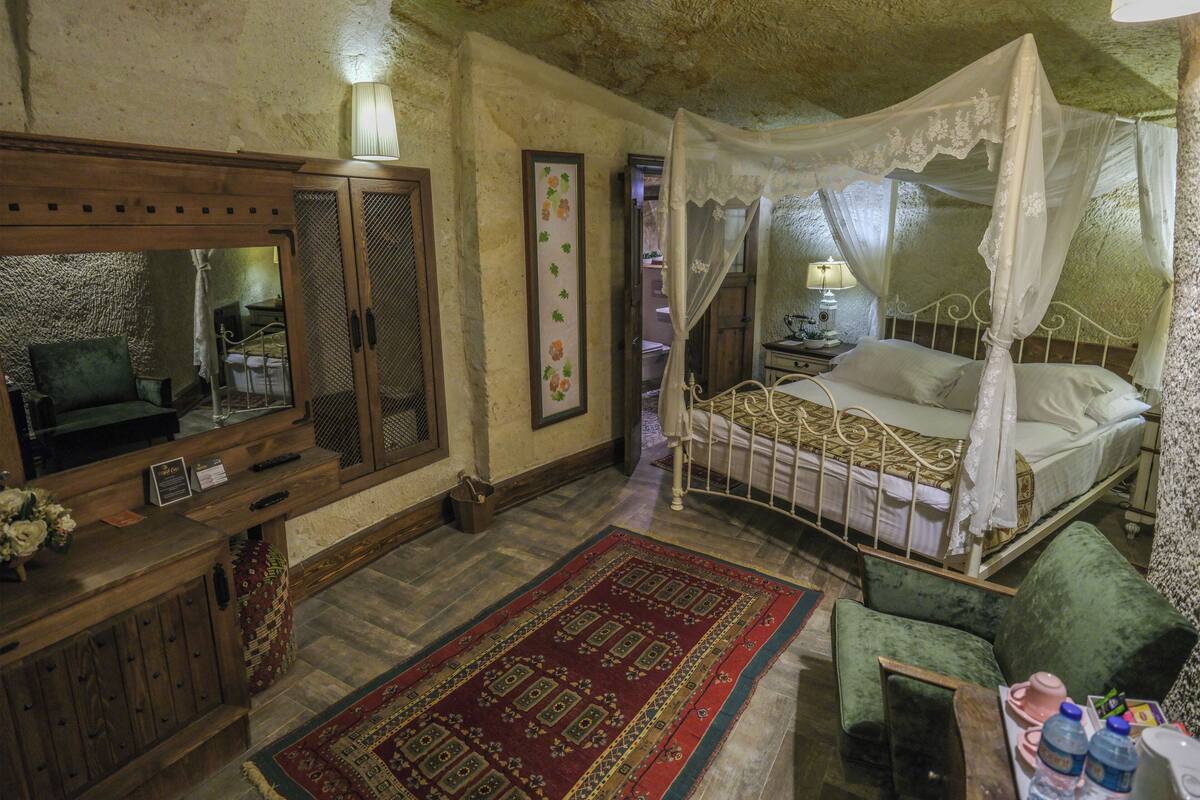 Un dormitorio con cama con dosel, un aparador de madera, un espejo, un sillón verde y una alfombra roja con estampado.