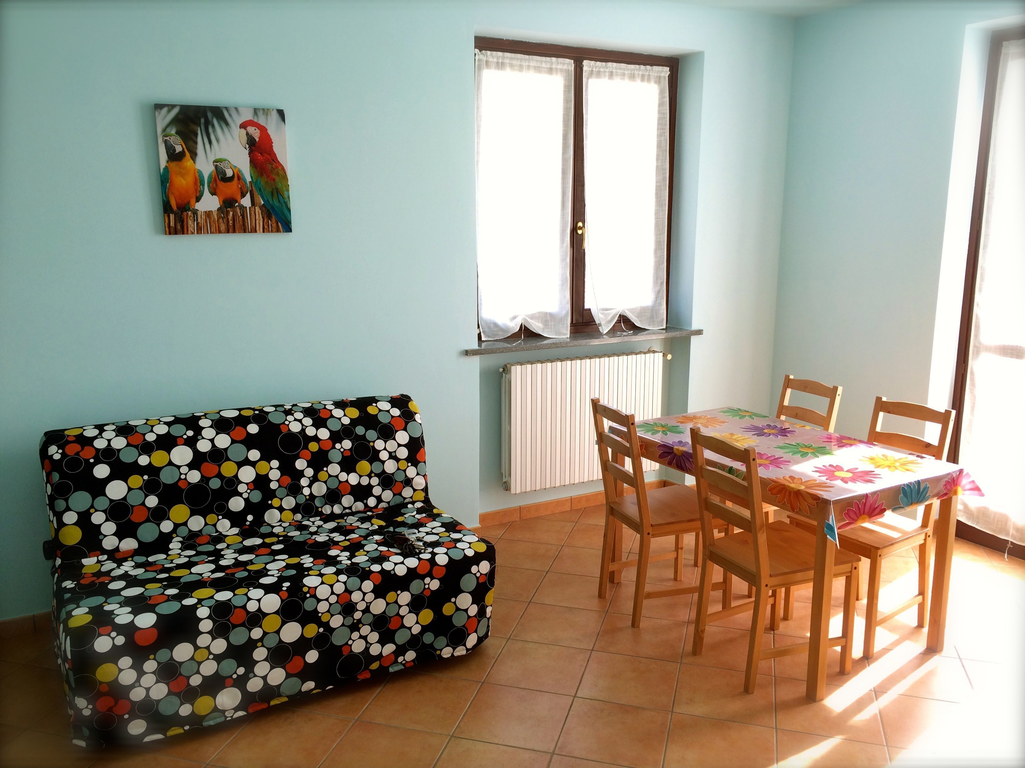 A room with a polka-dotted sofa, a dining table with chairs, a window with curtains, and a radiator.