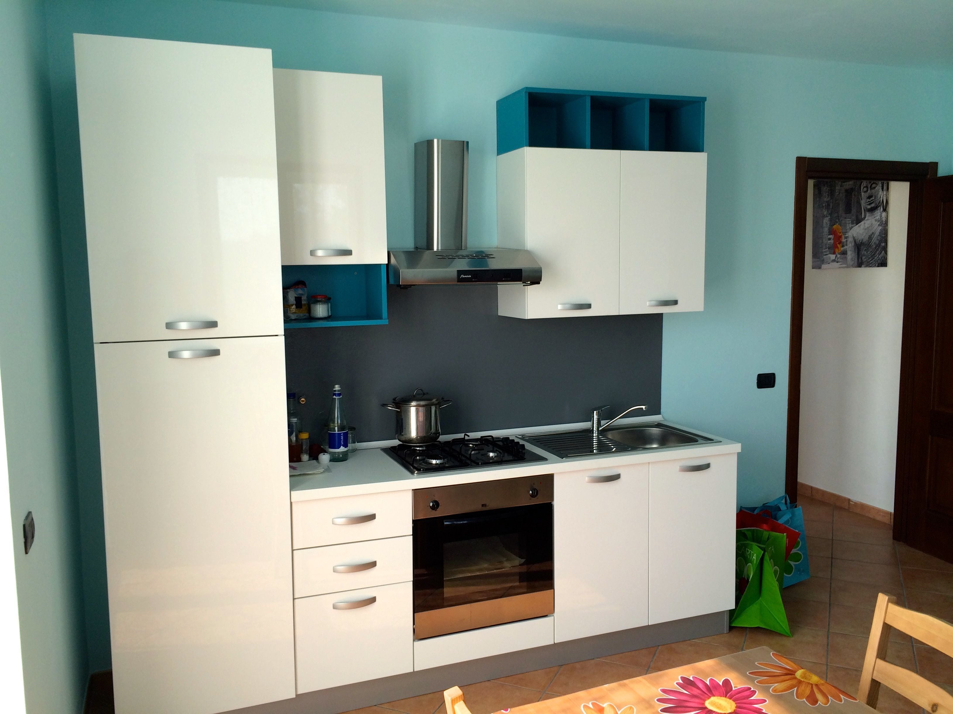 A compact kitchen with white cabinets, a stove, oven, and a sink. There are blue storage shelves above the stove and a small window on the right.