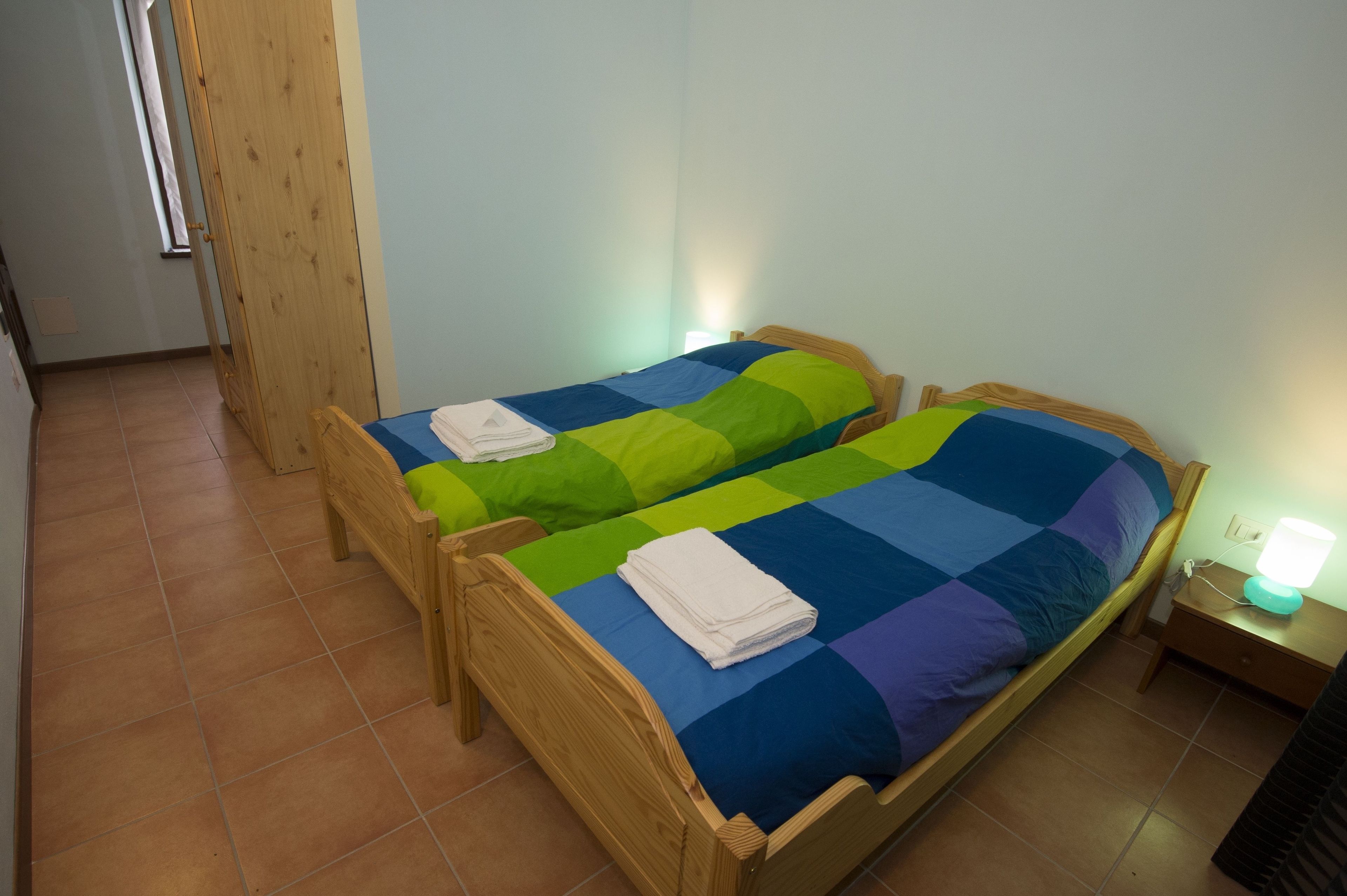Una stanza con due letti di legno, ognuno con una coperta colorata. Sul lato destro, un comodino con una lampada.
