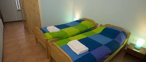 Una habitación con dos camas de madera, cada una con una colcha colorida. A la derecha, un mesita de noche con una lámpara.