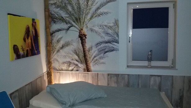 A bedroom with a bed, a wall-mounted window, and a wall art depicting a palm tree.