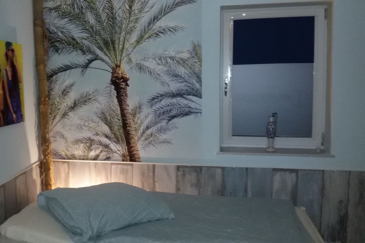 A bedroom with a bed, a wall-mounted window, and a wall art depicting a palm tree.
