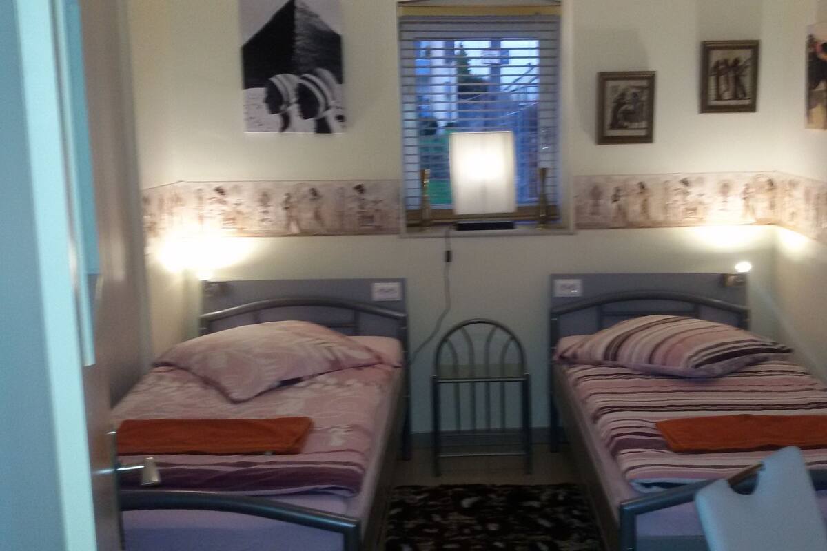 A narrow room with two single beds, a window with blinds, and wall-mounted artwork.
