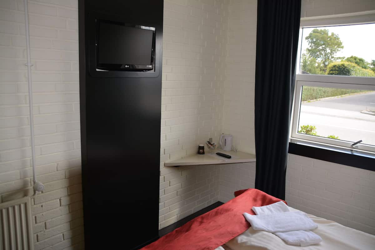 A hotel room with a bed, a TV mounted on a black wall, a window with a view of the street, and a small shelf with a kettle and remote control.