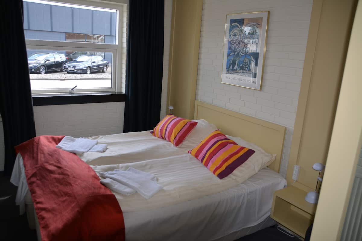 A hotel room with two beds, a window with a view of parked cars, a framed picture on the wall, and a bedside table with a lamp.
