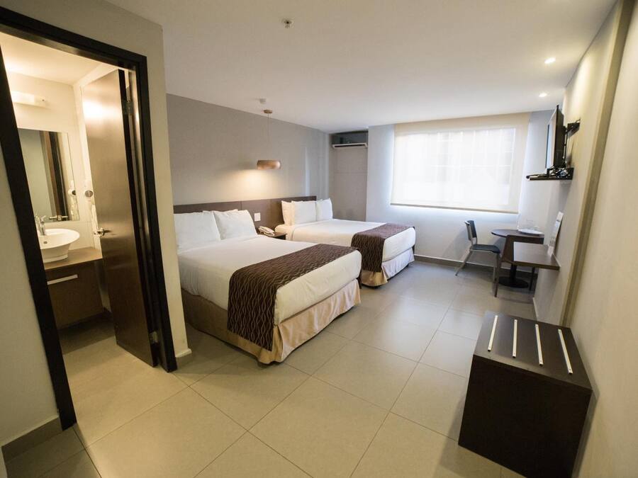 A hotel room with two beds, a desk, a chair, a small table, a mirror, and a window.