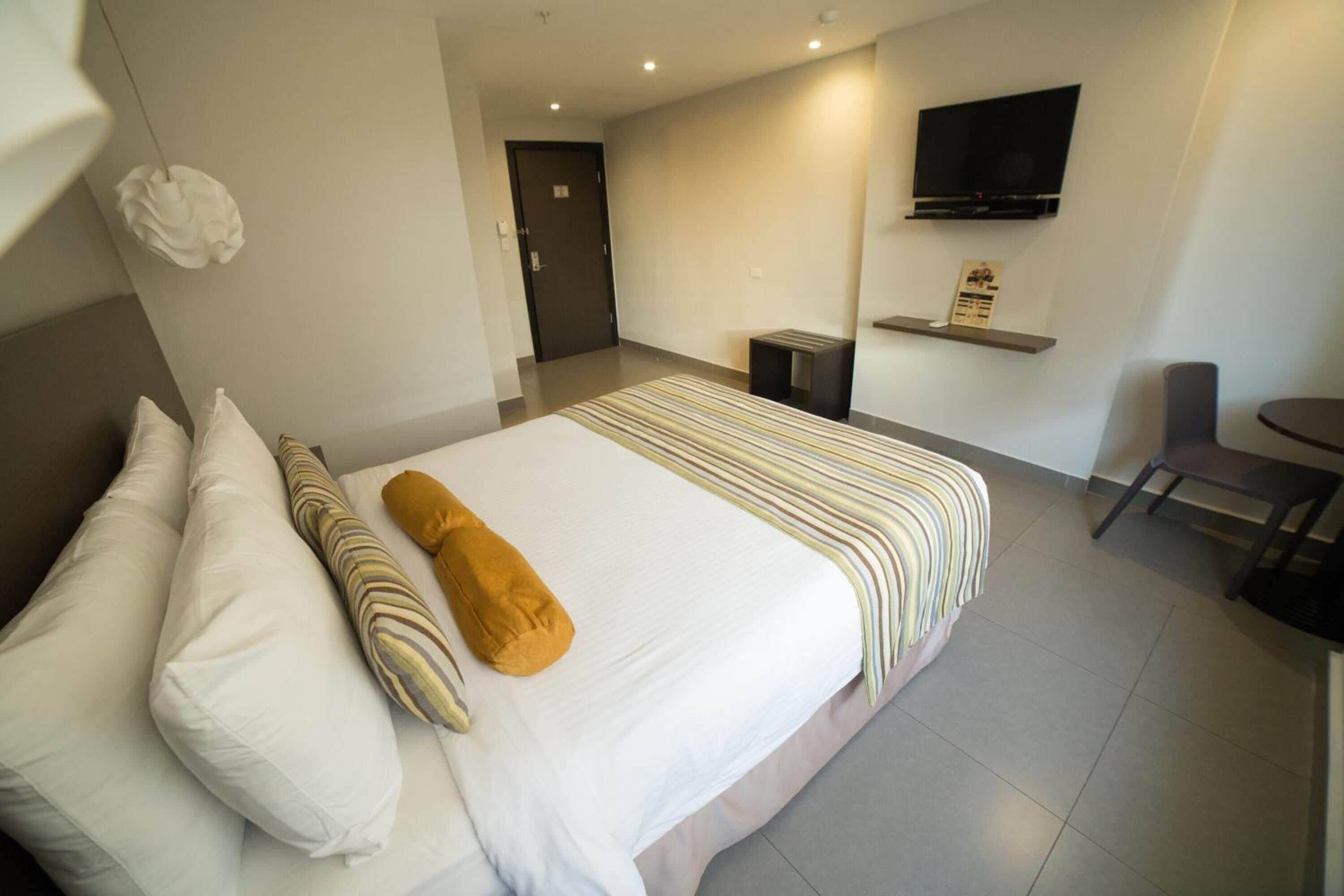 A hotel room with a bed, pillows, a television, a chair, and a small table.