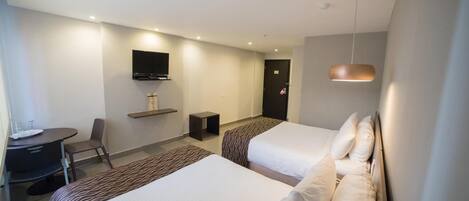 A hotel room with two beds, a desk with a chair, a TV mounted on the wall, and a small table.
