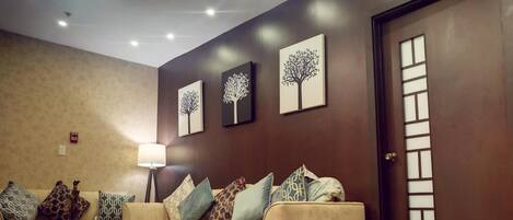 A living room with a beige sofa, patterned cushions, a glass coffee table, and a floor lamp. There are three framed artworks on the wall and a door with a frosted glass panel.