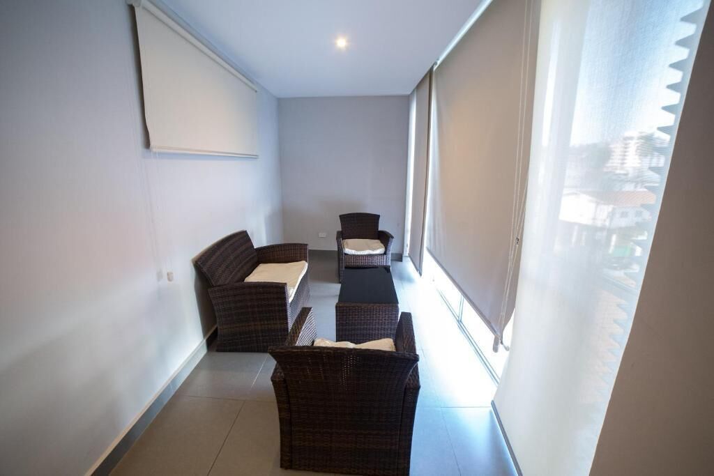 A narrow room with two wicker sofas and a small table, large windows with blinds, and a white ceiling.