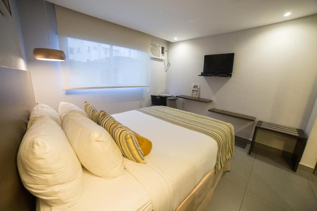 A hotel room with a large bed, a desk, a TV, and a window with blinds.