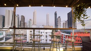 A rooftop bar with four metal bar stools and a wicker chair, overlooking a cityscape with high-rise buildings. - Hotel Ojos del Rio (Panama City)