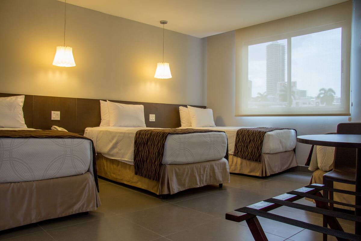 A hotel room with two beds, a small table, and a window with a city view.