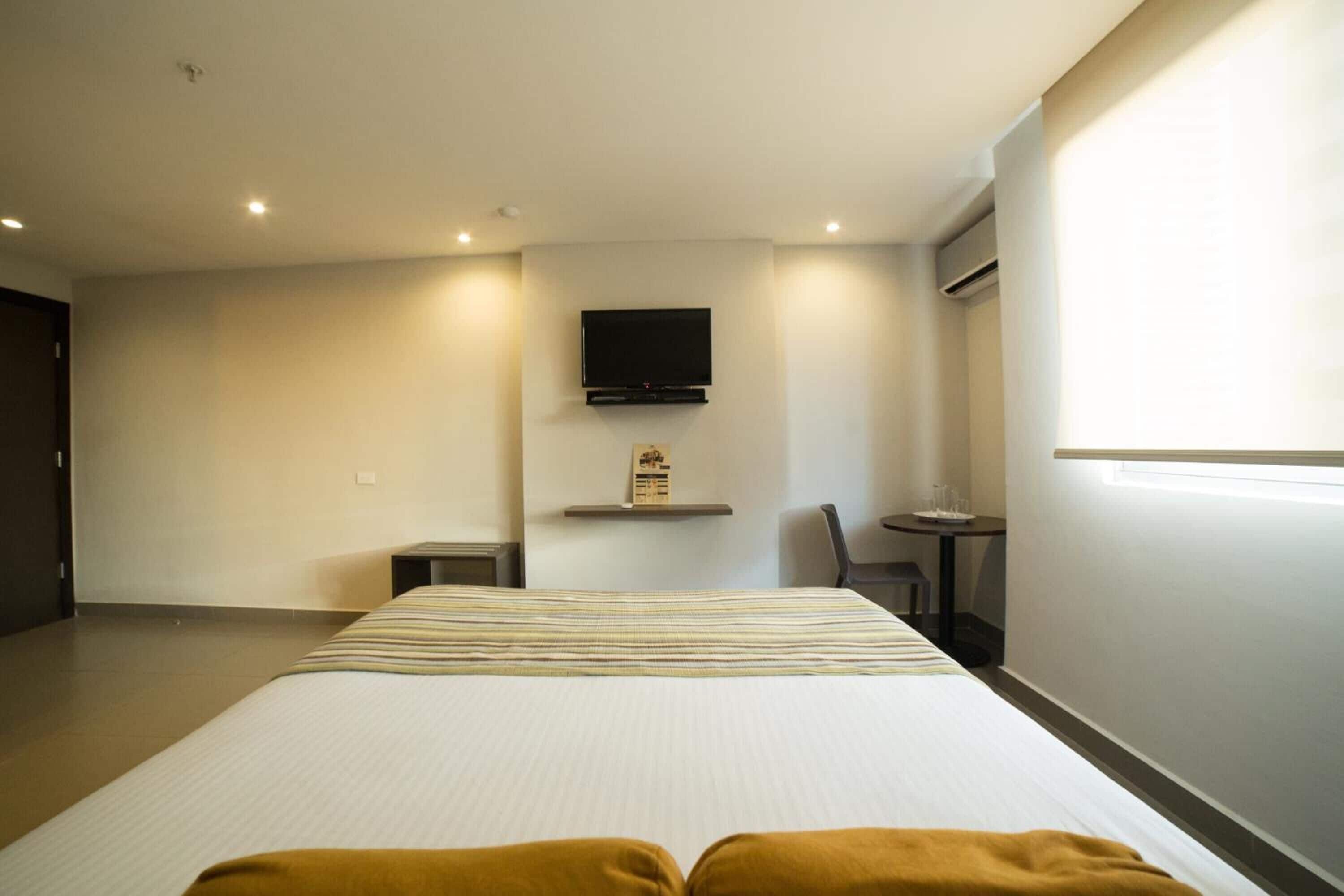Habitación de hotel con una cama grande, un televisor montado en la pared, una mesita pequeña y una silla.