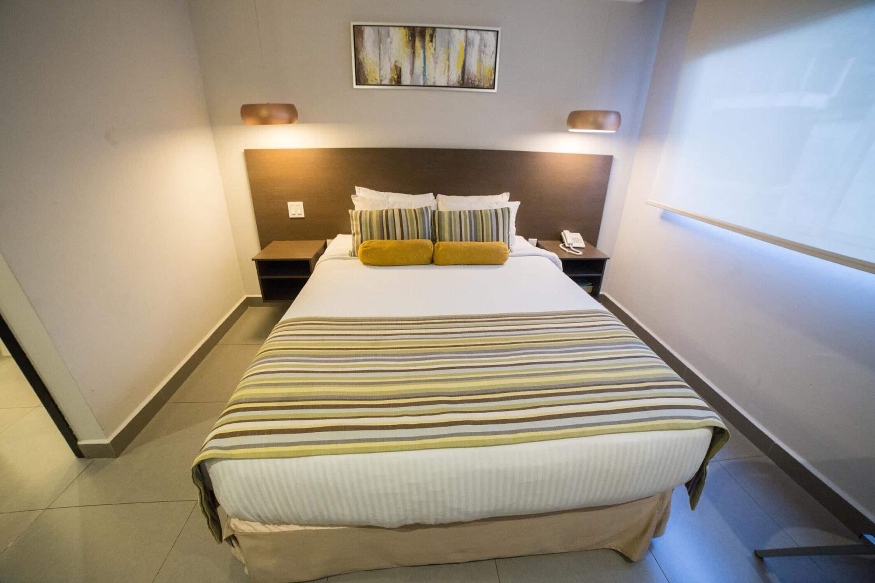 A hotel room with a large bed, a headboard, two bedside tables, a nightstand, a wall-mounted projector, and a window.