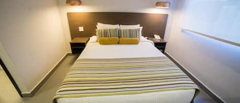 A hotel room with a large bed, a headboard, two bedside tables, a nightstand, a wall-mounted projector, and a window.