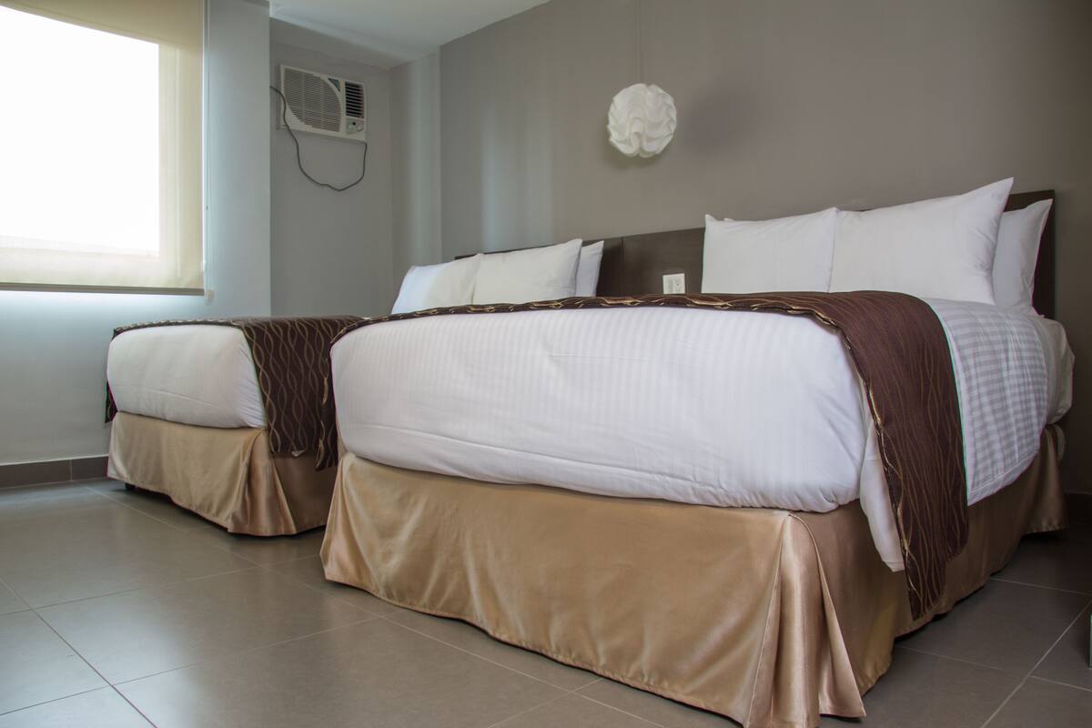 A hotel room with two beds, beige bedding, a brown throw blanket, a white wall decoration, and an air conditioning unit.
