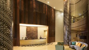Zona de recepción con un mostrador de madera, dos empleados, un sillón verde, un sofá gris con cojines de colores y una alfombra estampada.