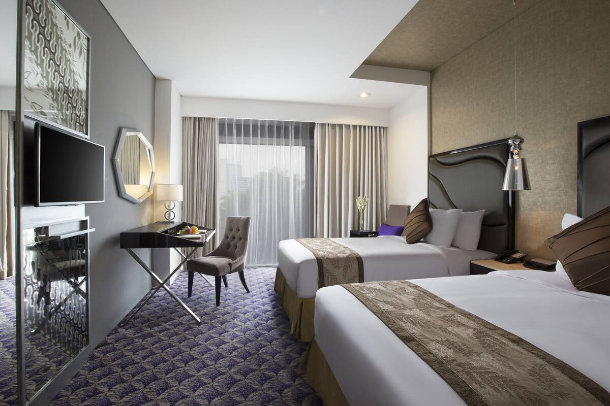 Habitación de hotel con dos camas, un escritorio con una silla, un televisor y un ventanal con cortinas.