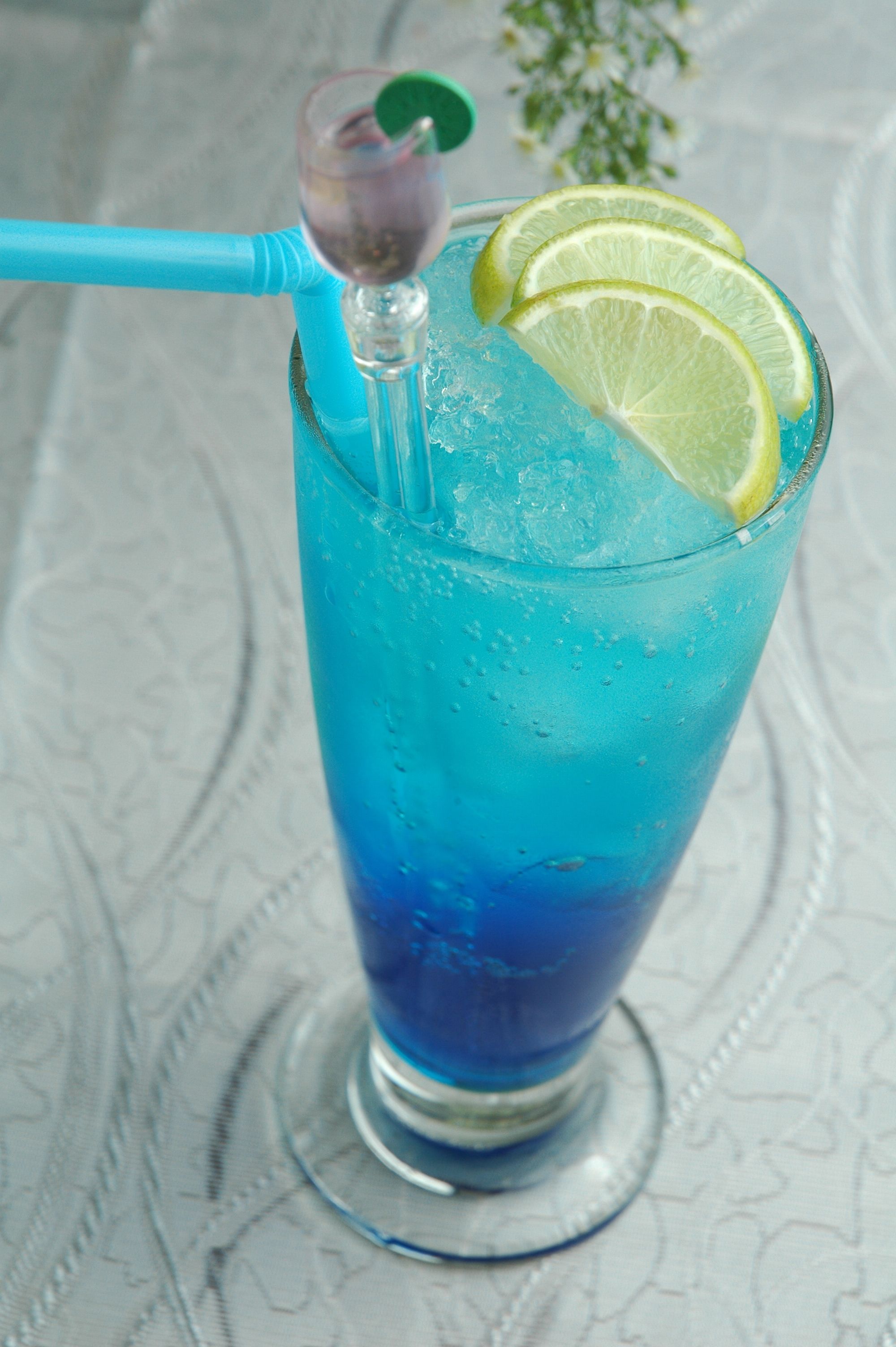 Un vaso alto con una bebida azul, hielo y una rodaja de lima, con un popote azul.