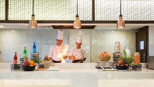 Uma área de cozinha com chefs preparando alimentos, um buffet com pratos e itens alimentícios, e uma parede decorativa com padrão geométrico.
