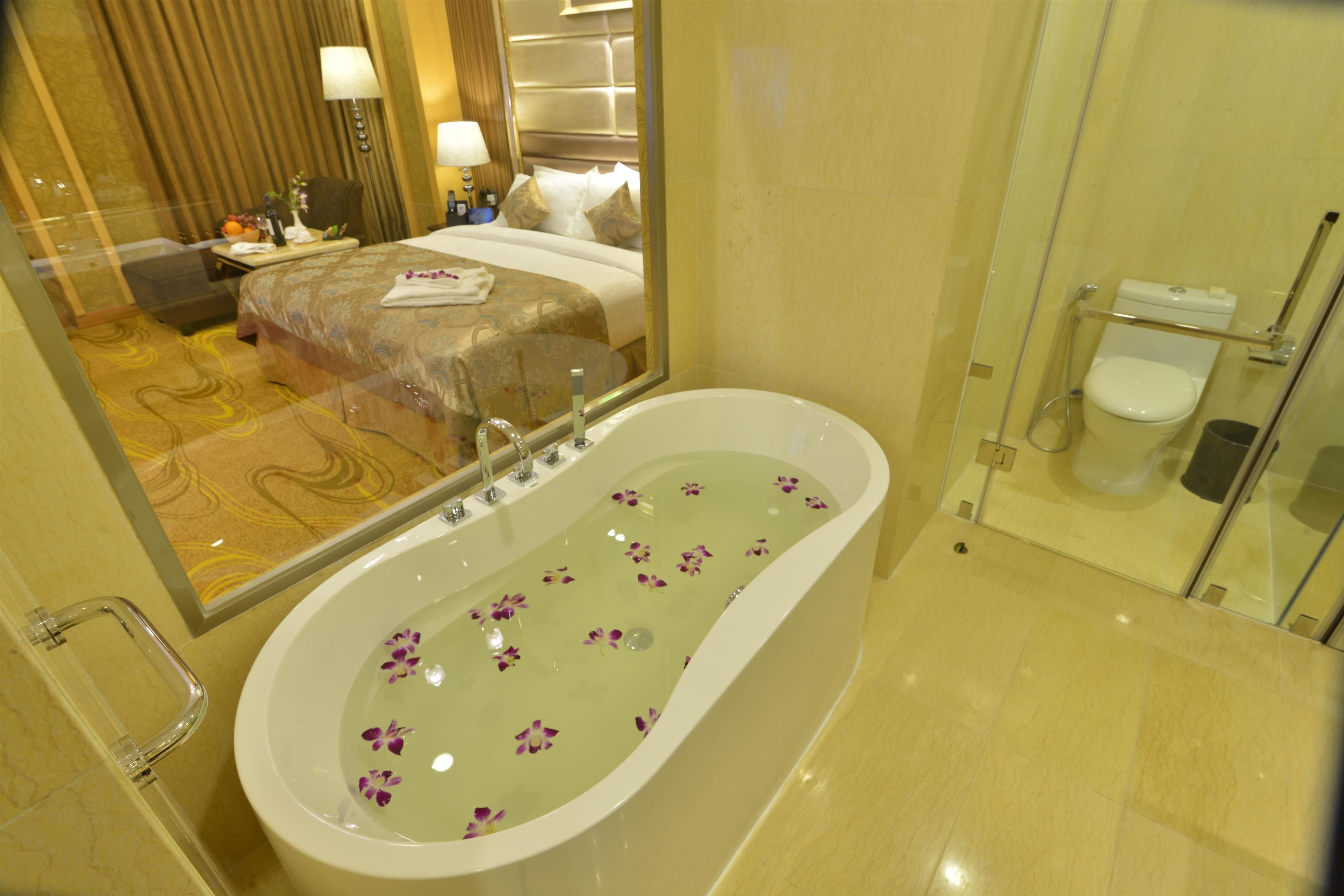A modern bathroom with a large bathtub, a glass shower enclosure, and a visible bed with a nightstand in the background.
