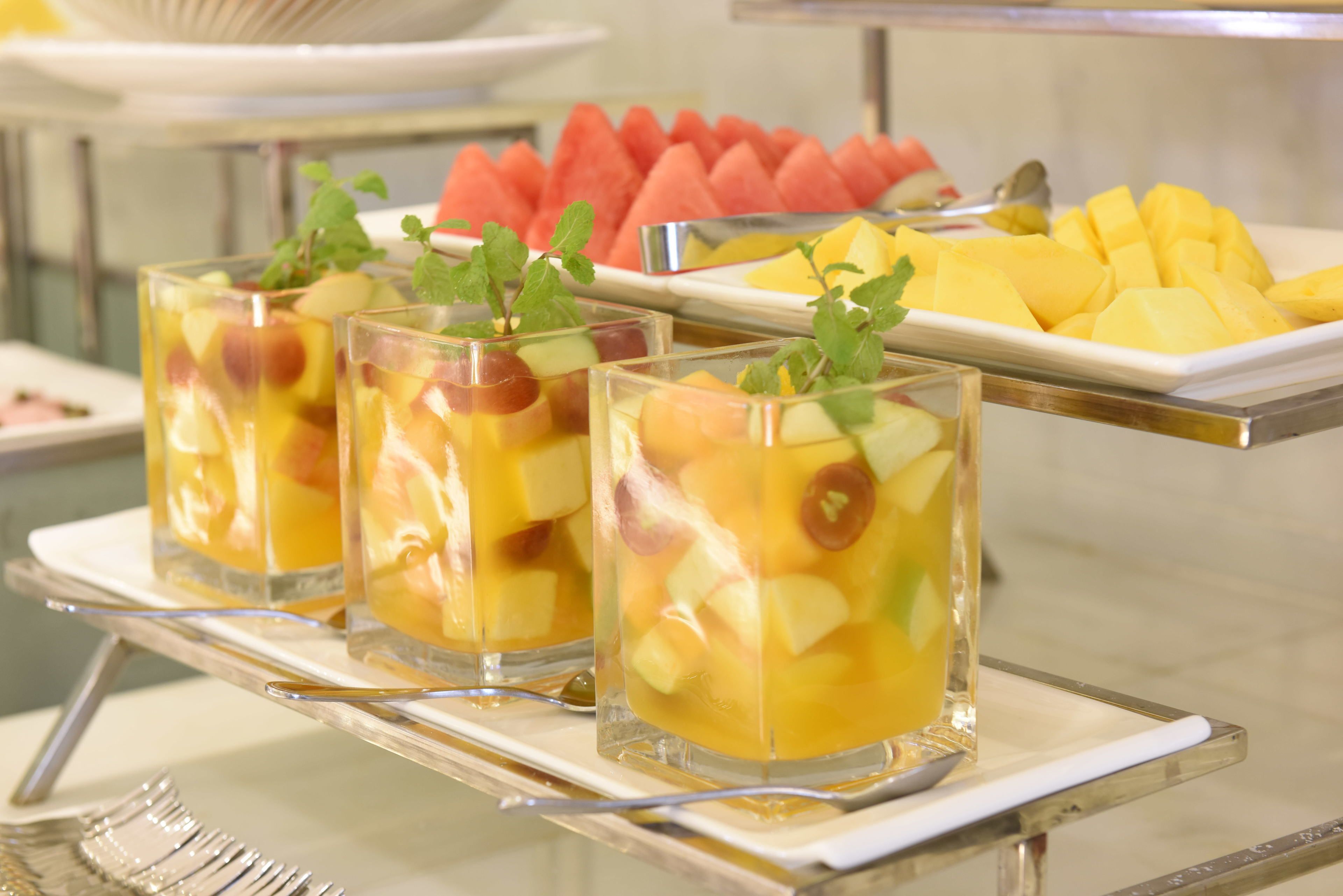 Three glasses of fruit punch on a tray with a spoon. Behind the tray, there are plates of watermelon and mango.