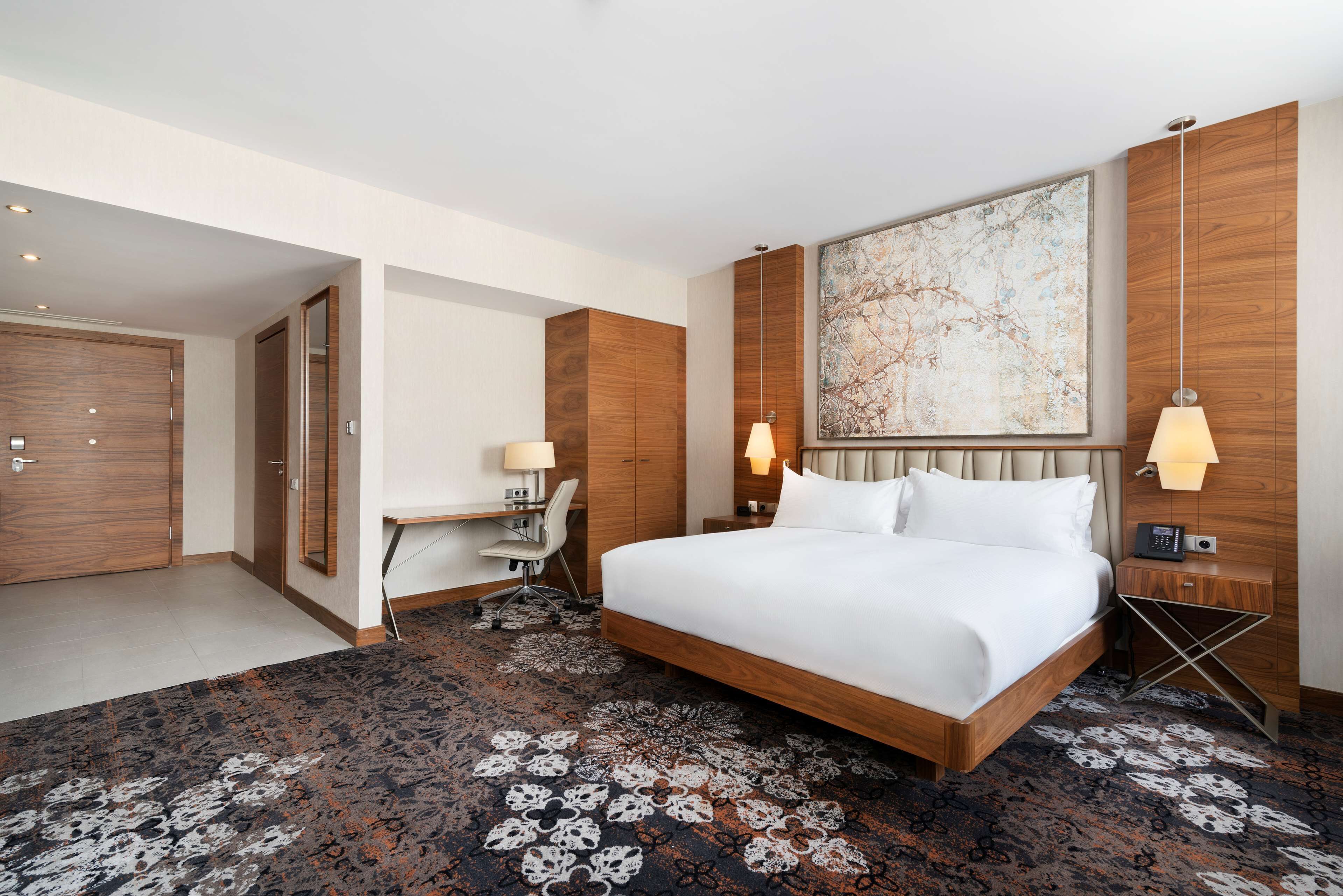 A hotel room with a large bed, a desk, and a chair, a wooden wardrobe, and a large abstract painting on the wall.