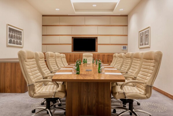 Una sala de conferencias con una larga mesa de madera, sillas de cuero color beige, un televisor de pantalla plana y cuadros en las paredes.