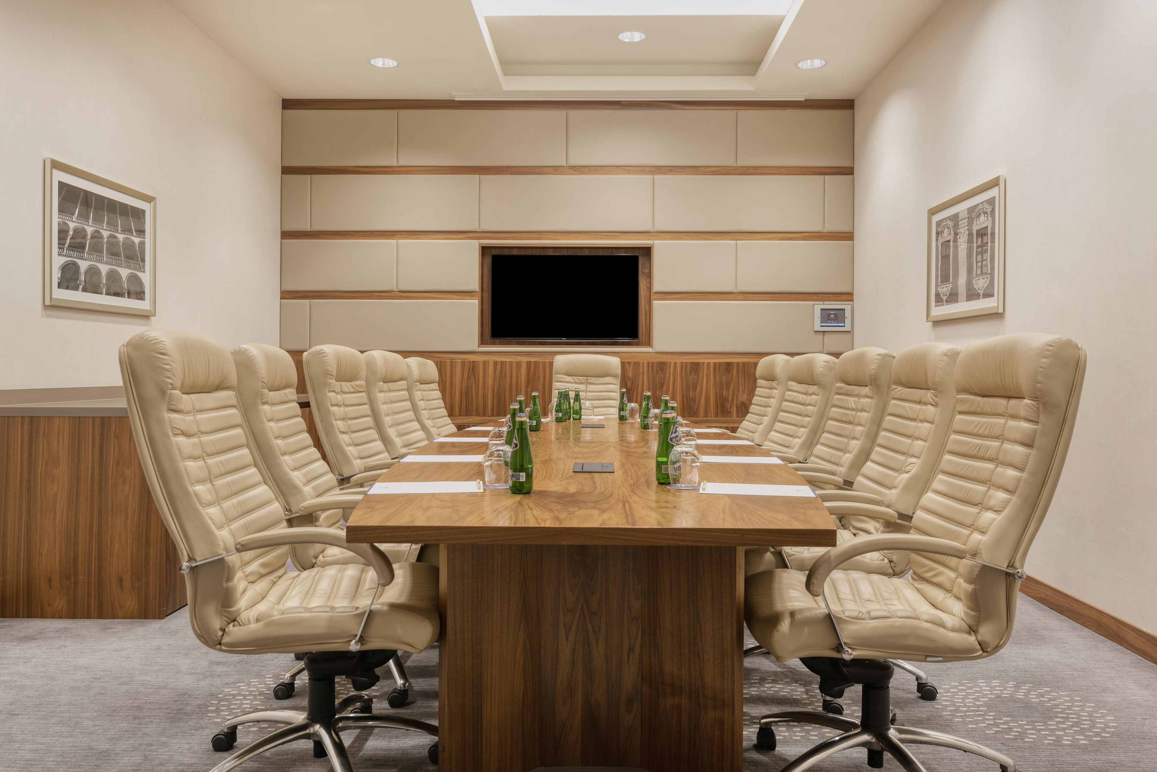 A conference room with a large table, beige leather chairs, a flat-screen TV, and framed pictures on the walls.