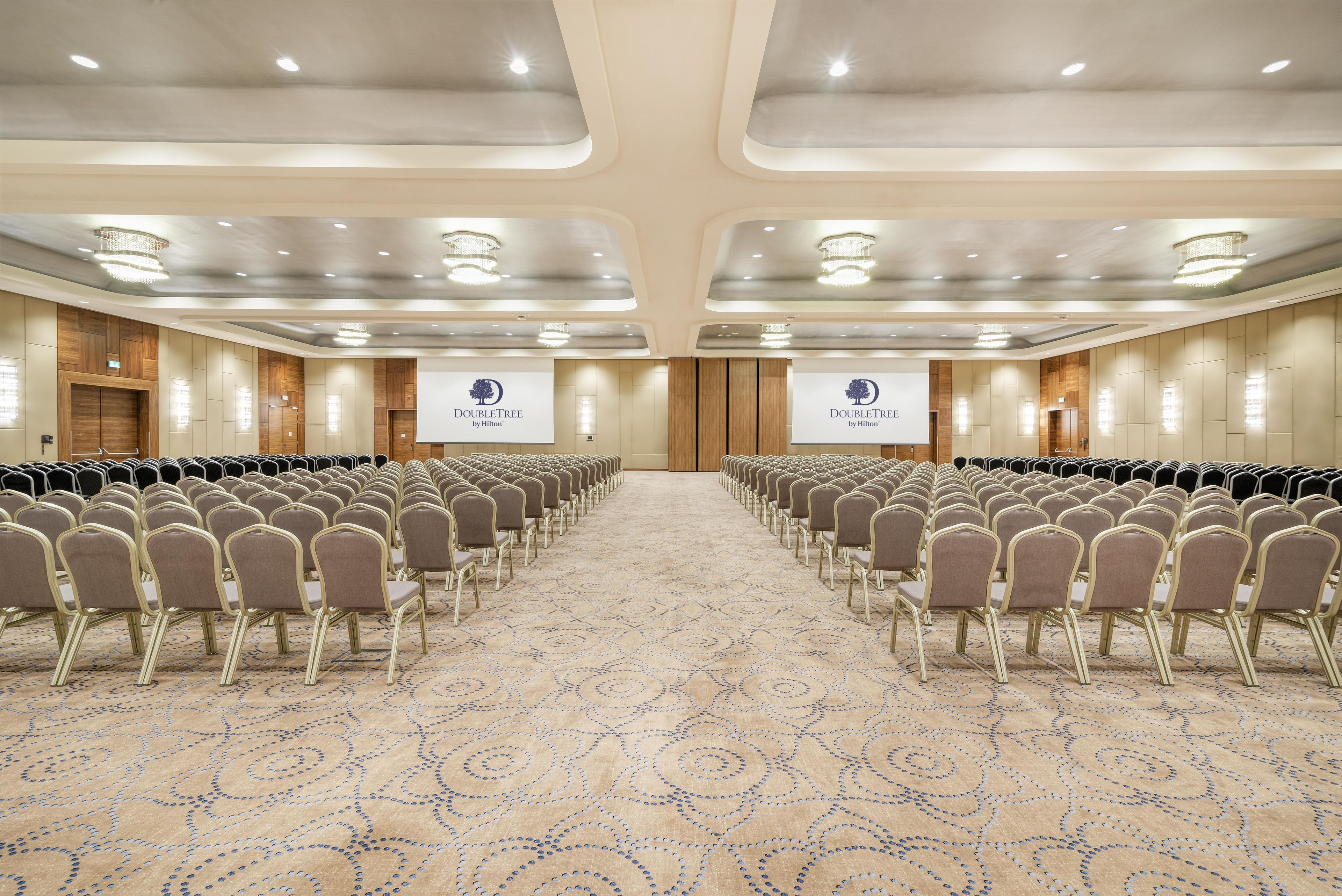 Un salón de conferencias con filas de sillas, piso alfombrado e iluminación en el techo.