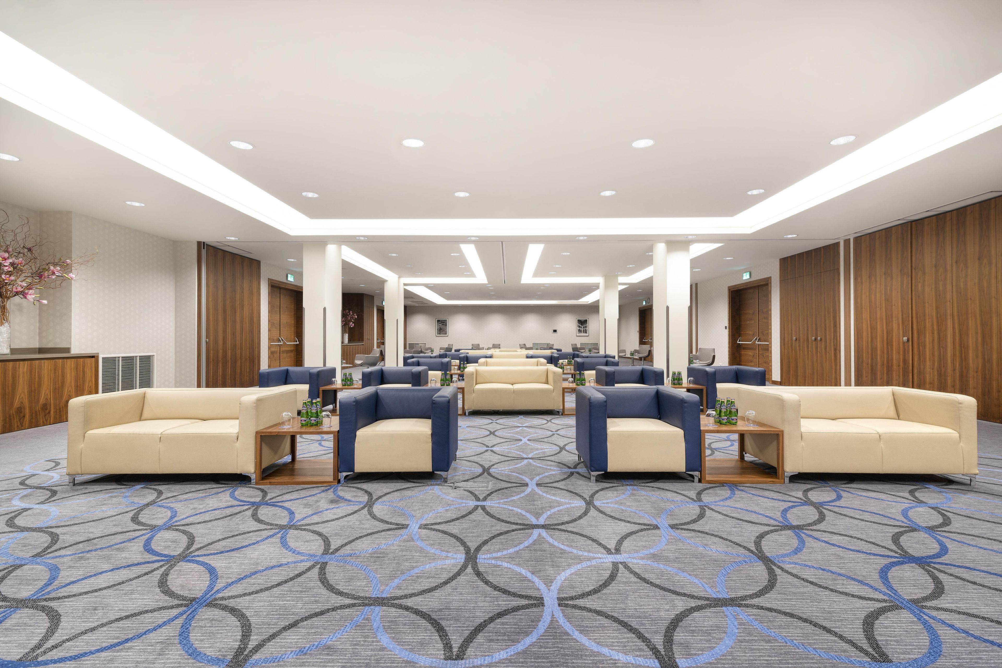 A hotel lobby with multiple seating areas, a patterned carpet, and a modern ceiling design.