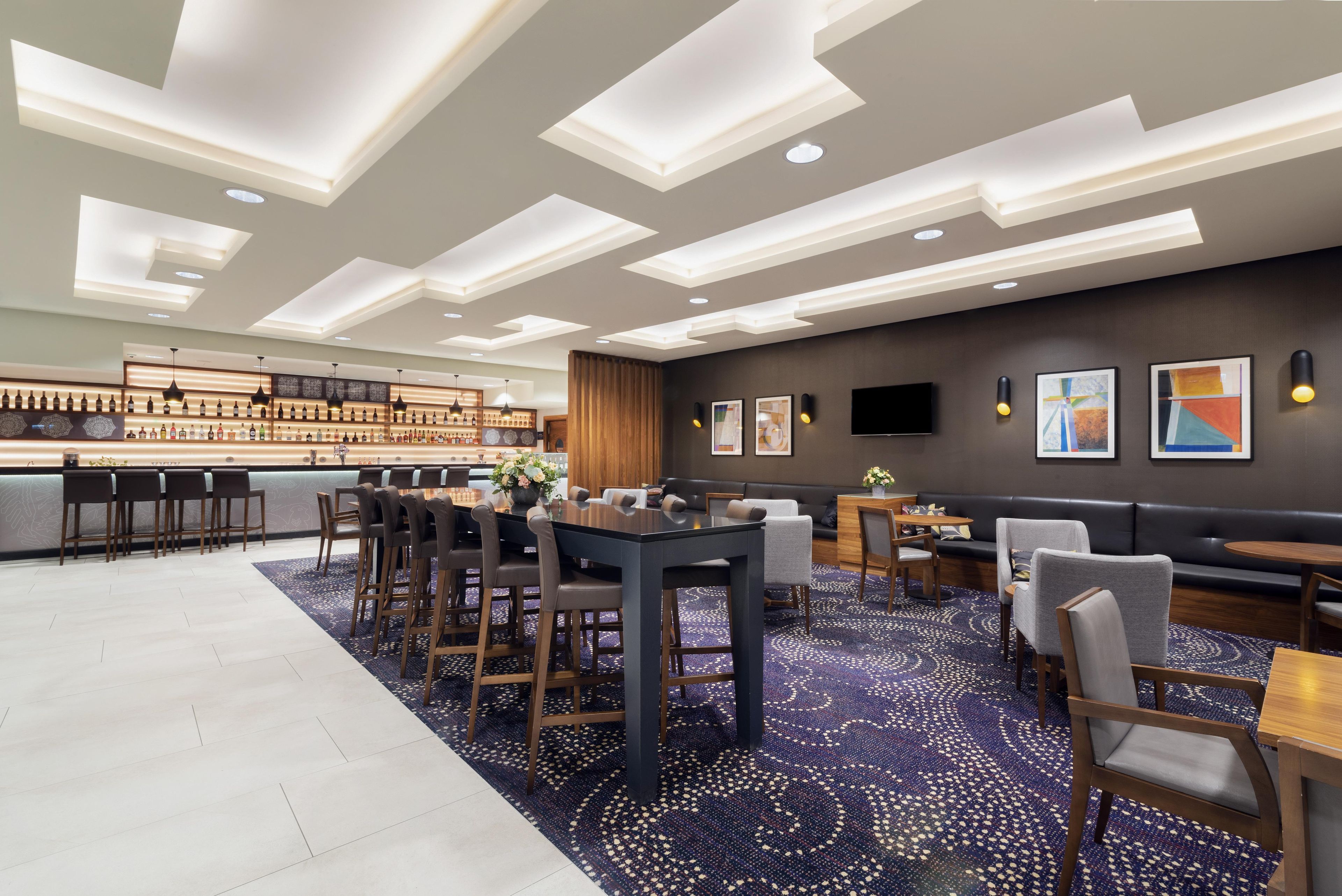 Zona de bar moderna con una barra alargada, varios taburetes y un área de estar con sofás y sillones.