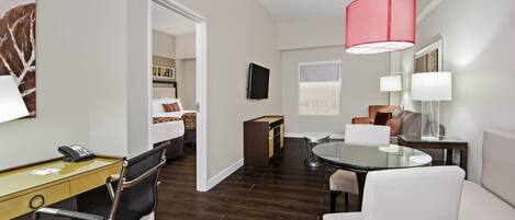 A hotel room with a desk, chair, round table, and a bed in the background.