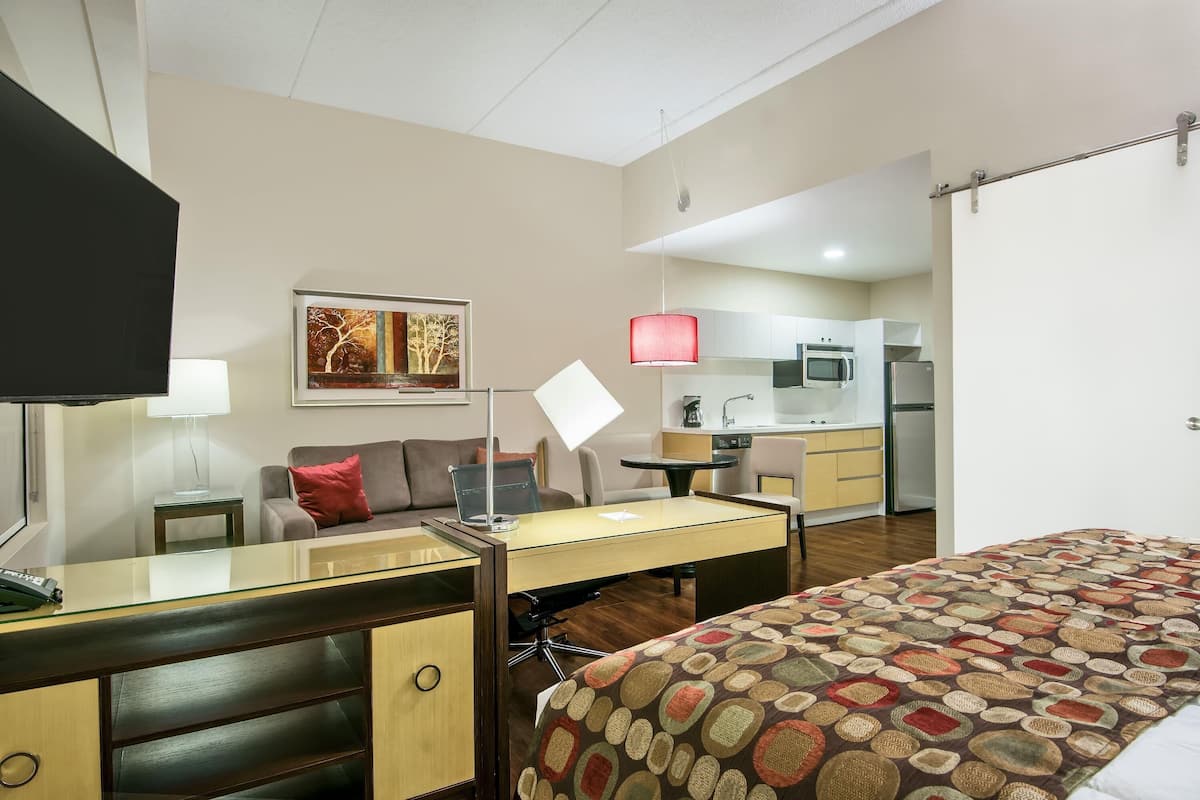 A hotel room with a bed, desk, chair, lamp, artwork, and a kitchenette.