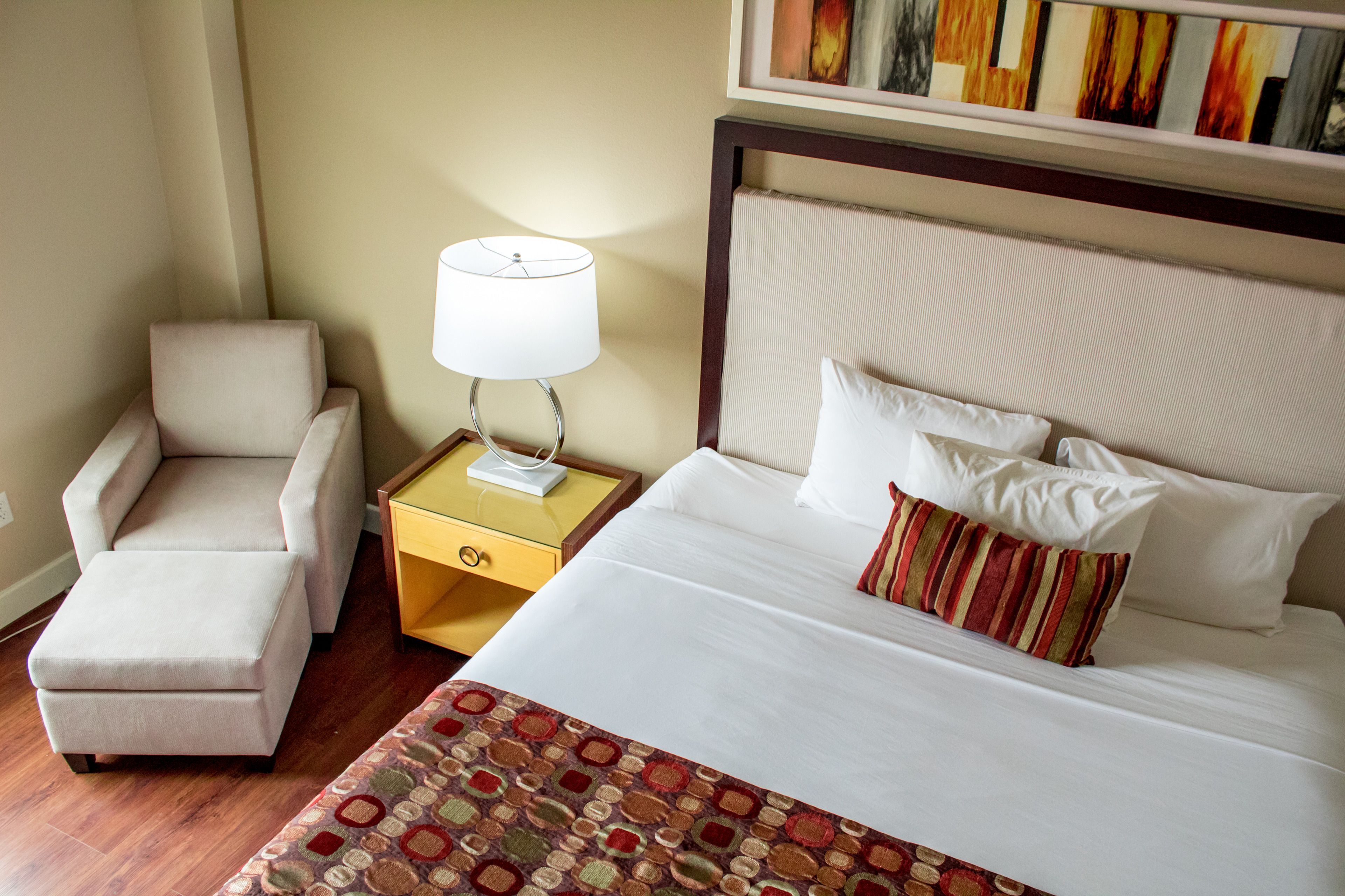 A hotel room with a bed, a chair, a nightstand with a lamp, and a painting on the wall.
