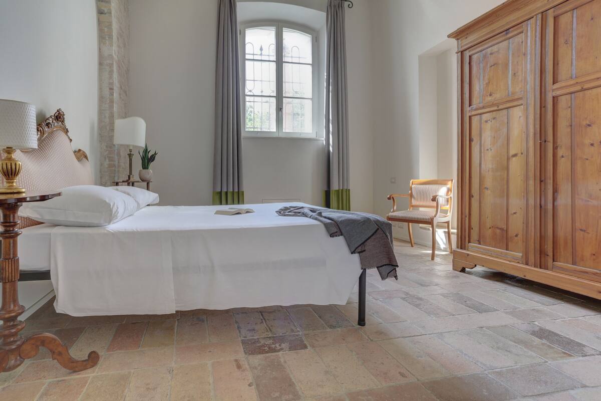 Un lit bien fait, recouvert d’une couvre-lit blanche, une table de chevet avec une lampe, une fenêtre avec des rideaux et une armoire en bois.
