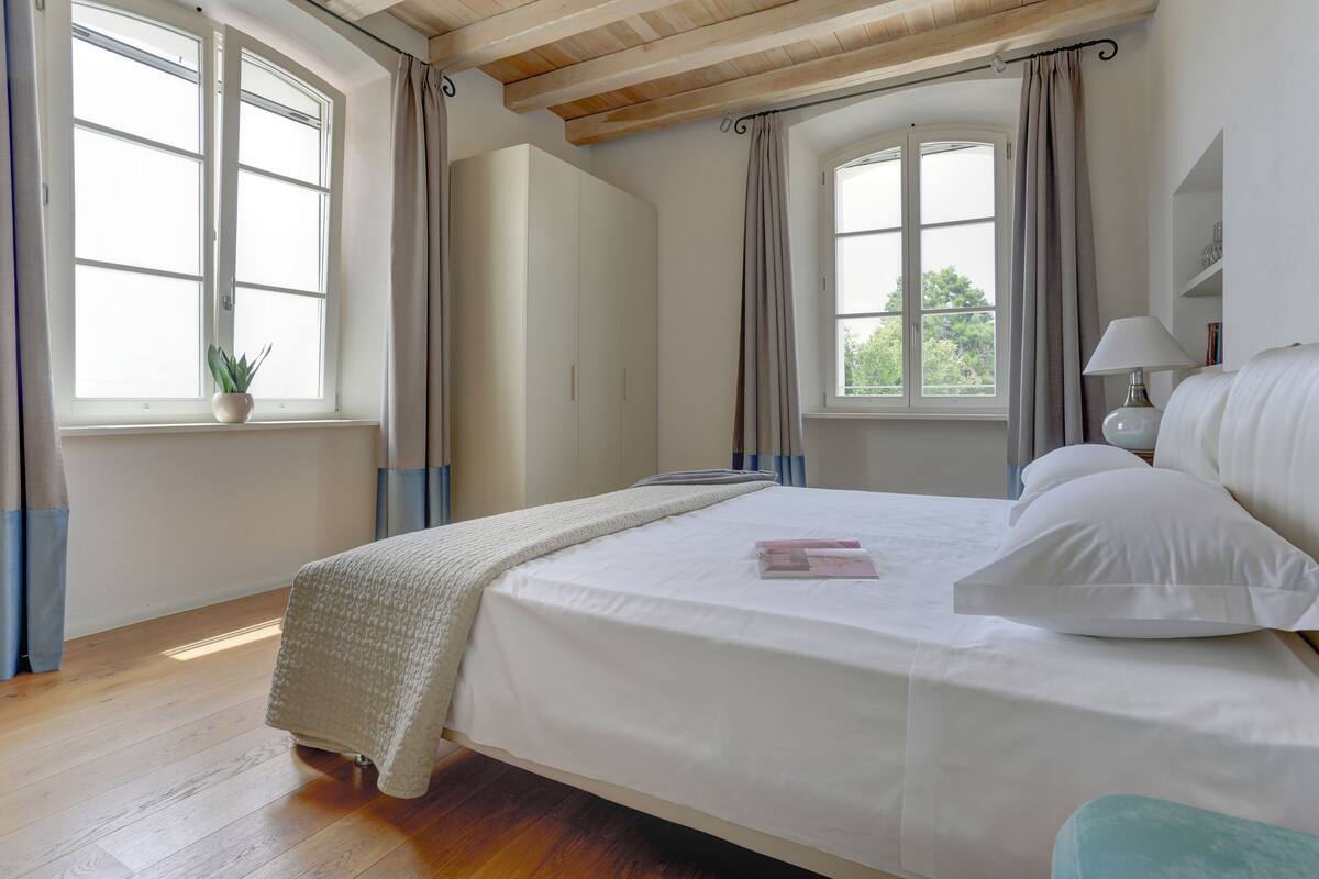 Une chambre à coucher avec un grand lit, une table de chevet, une lampe et une fenêtre avec des rideaux.