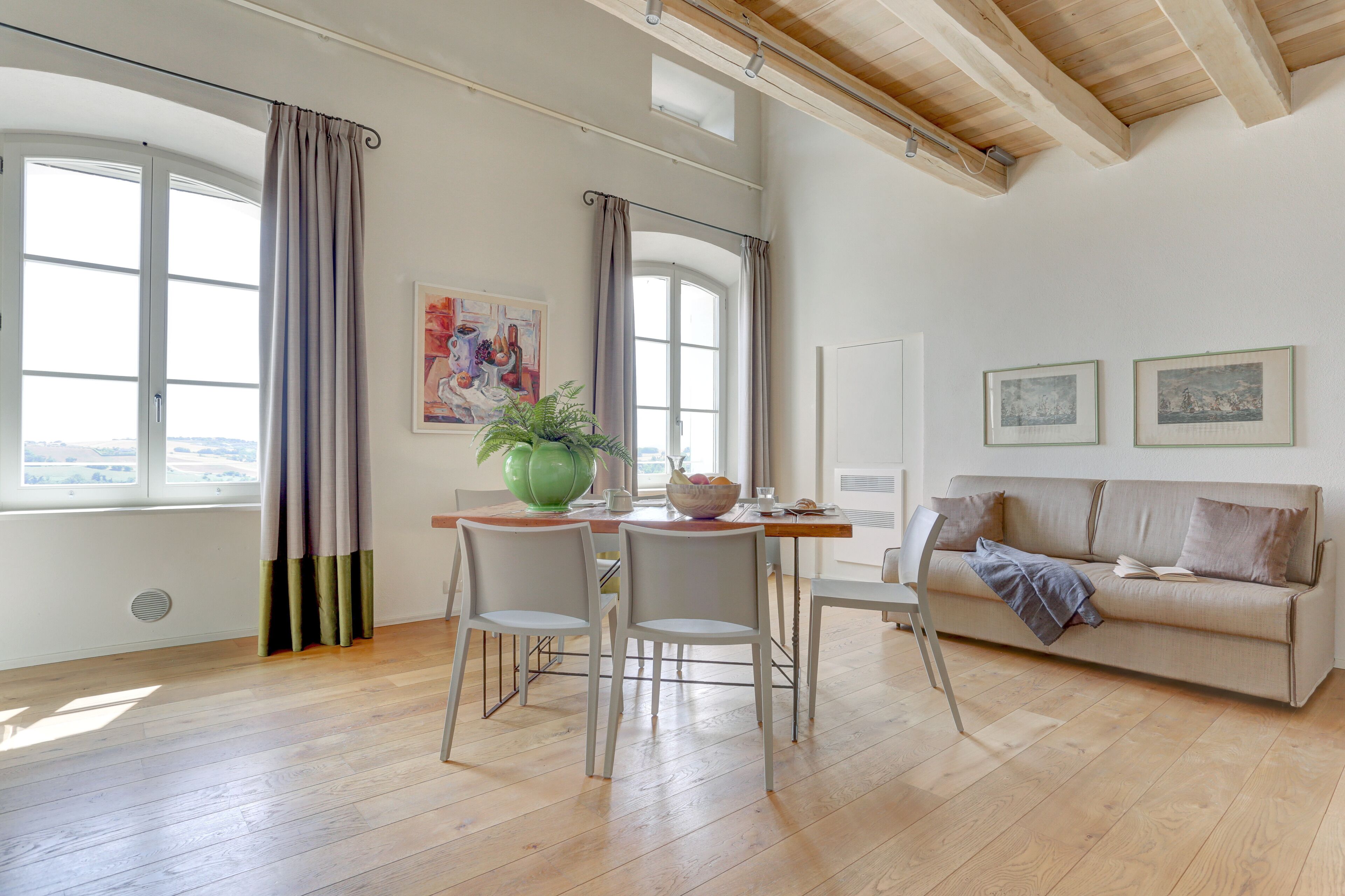 A spacious living area with a wooden floor, a dining table with chairs, a sofa, and a large window with curtains.