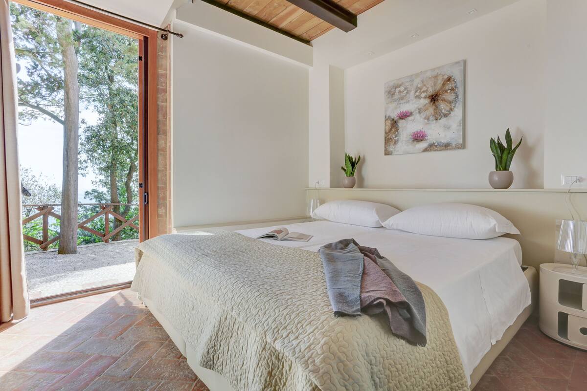 Une chambre avec un grand lit, un plafond en bois, un tableau accroché au mur et une vue sur les arbres à travers une porte ouverte.