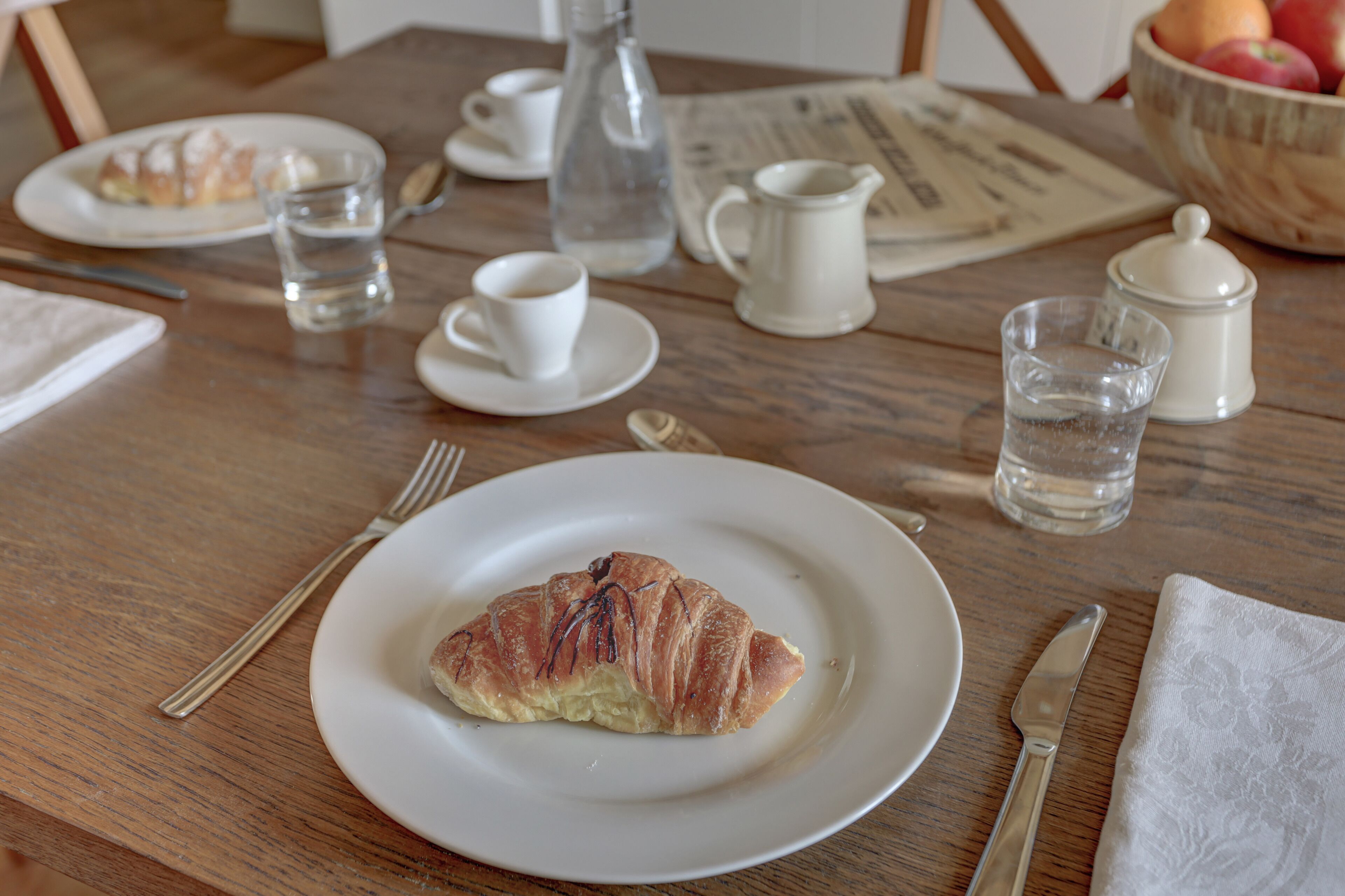 Egy reggelizőasztal, rajta egy croissant, egy kávéscsésze, egy pohár víz és egy újság.