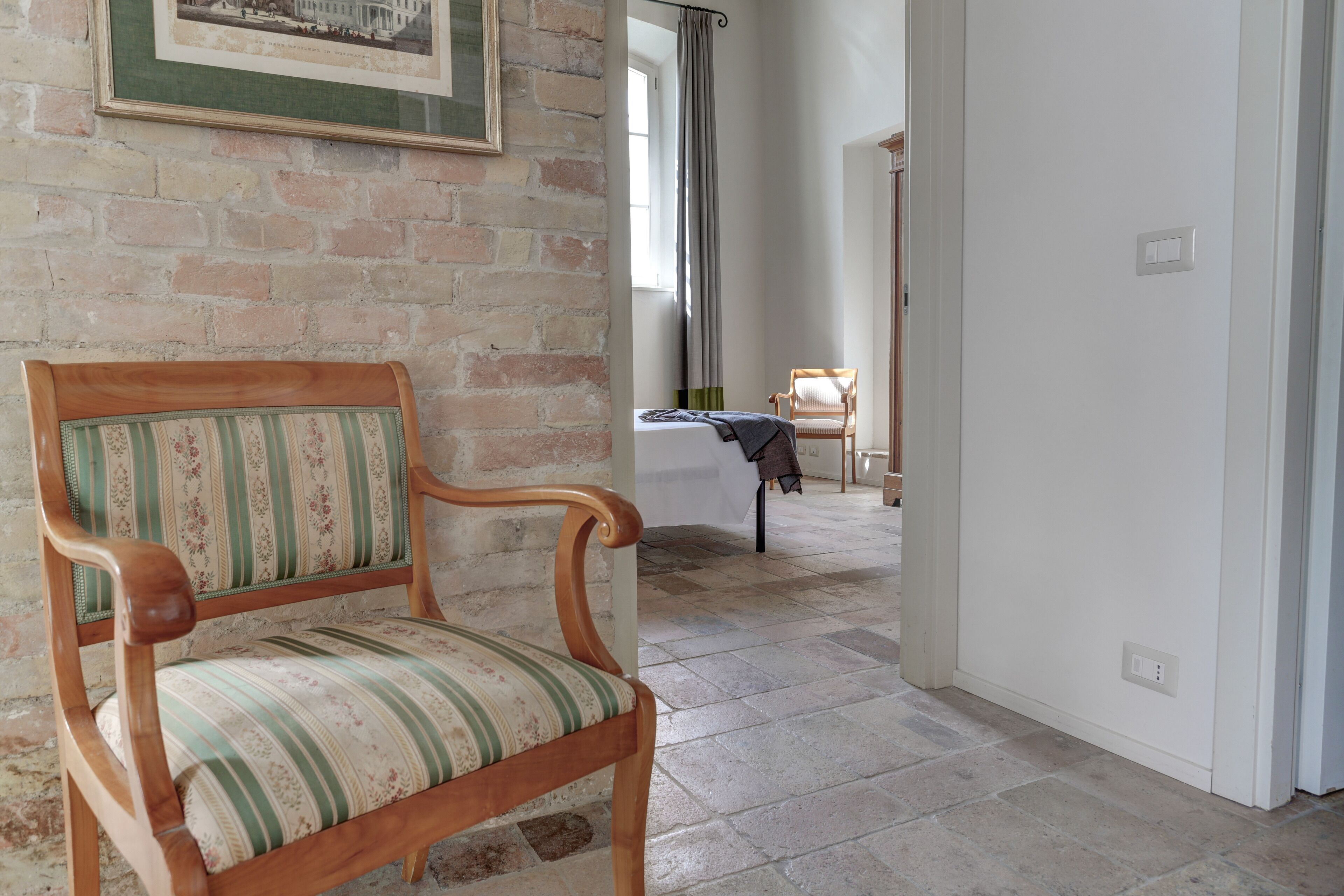 A room with a wooden chair with striped upholstery, a bed with a white cover, and a framed picture on the brick wall.