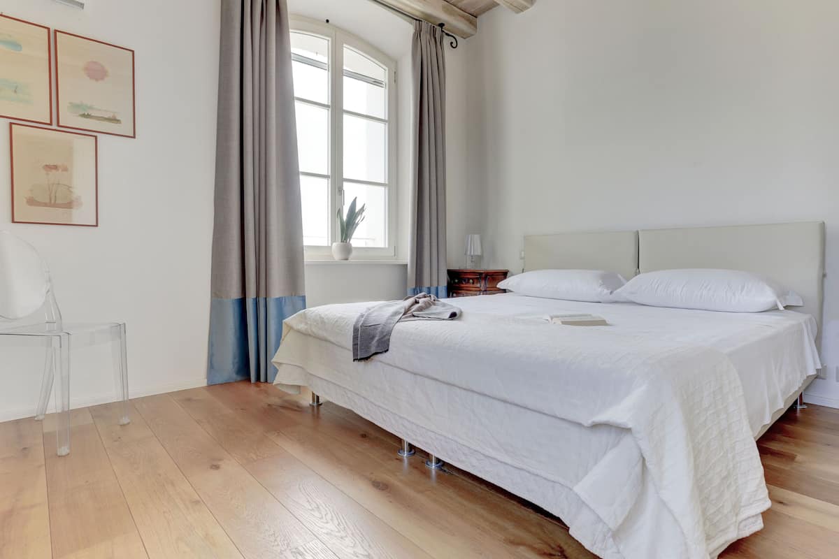 Une chambre avec un grand lit, une table de chevet, une chaise et une fenêtre avec des rideaux.