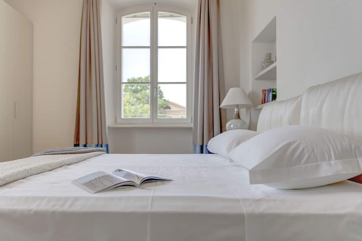 Un lit bien fait, avec un livre à portée de main, une fenêtre avec des rideaux et une lampe de chevet.