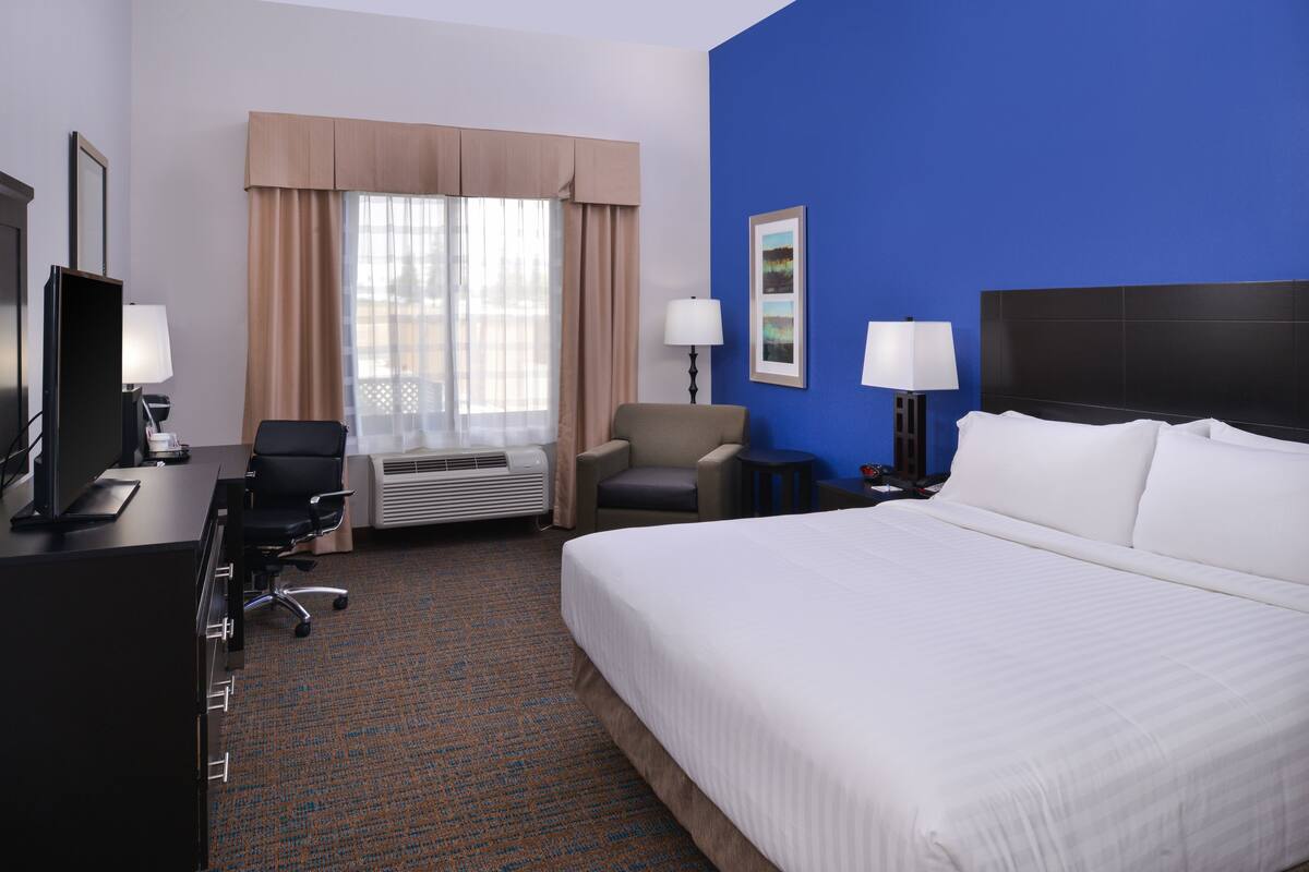 Ein Hotelzimmer mit einem großen Bett, einem Schreibtisch mit Computer, einem Sessel, einer Lampe und einem Fenster mit Vorhängen.