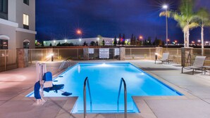 Une piscine dotée de deux rampes métalliques, un espace de détente avec des fauteuils et une vue sur un bâtiment en arrière-plan.